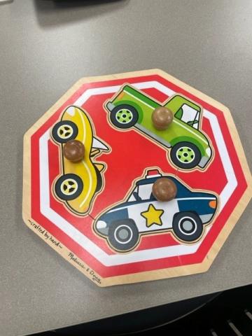 Wooden puzzle in the shape of a stop sign.  There are three cars, a yellow sports car, a green truck and a police car.  Each car has a large knob attached to allow for easy lifting