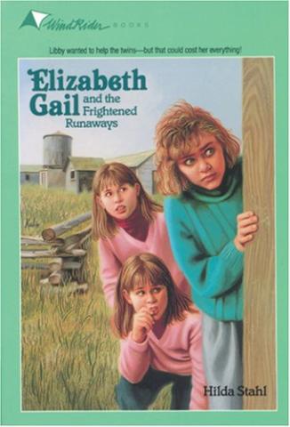 Three young girls peaking around a corner of a fence with a field in the background