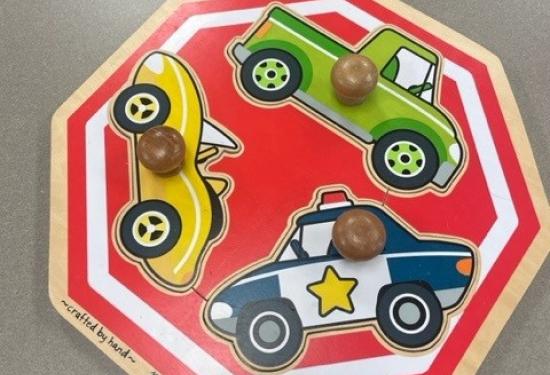 Wooden puzzle in the shape of a stop sign.  There are three cars, a yellow sports car, a green truck and a police car.  Each car has a large knob attached to allow for easy lifting