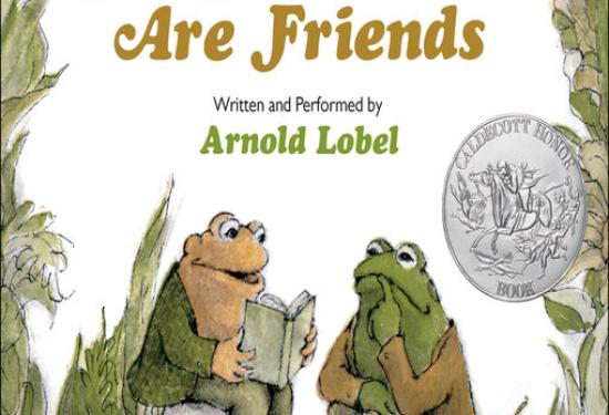 frog and toad sitting in grass with toad holding a book reading to frog