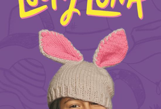 A girl with dark brown hair, smiling, with her hand on her left cheek, and wearing a knit cream hat with bunny ears